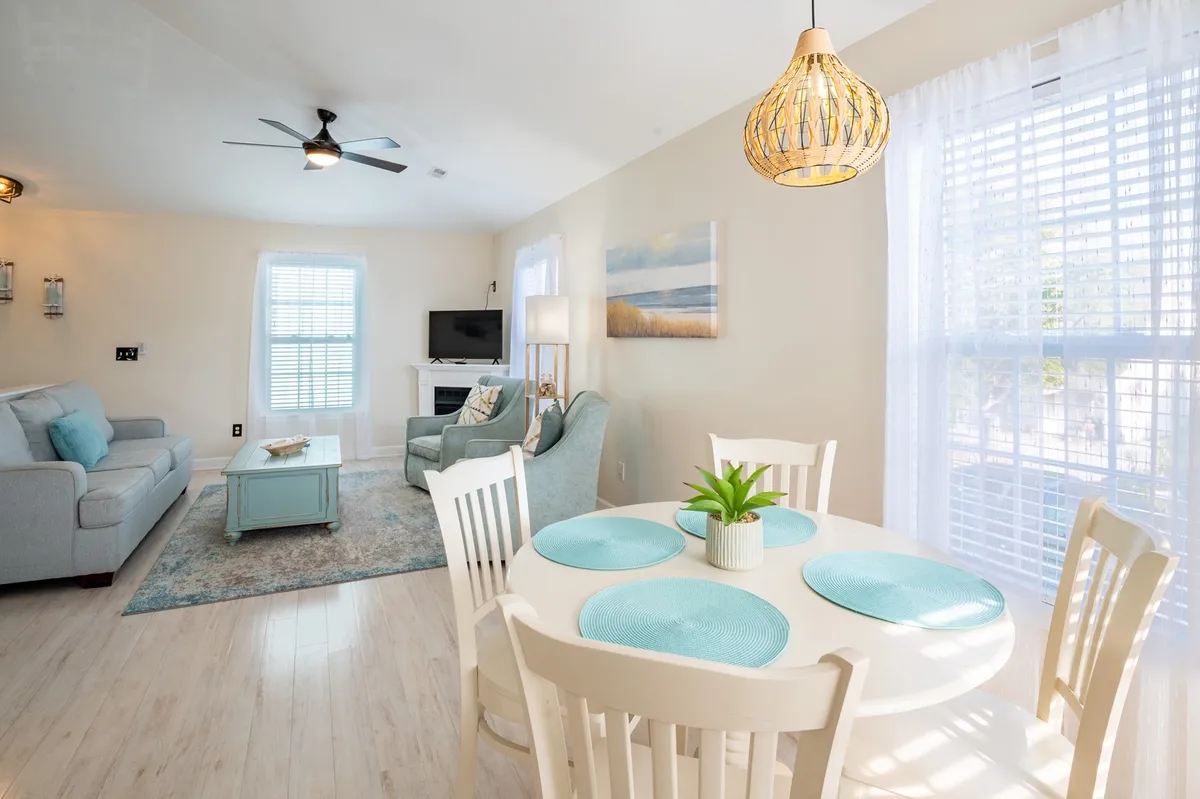 The image showcases a bright, cheerful living and dining area with a stylish light fixture above a welcoming dining table, a comfortable sofa for relaxing, and vibrant artwork on the wall. The space is perfect for gathering, sharing meals, and unwinding together after a sunny day at Carolina Beach.