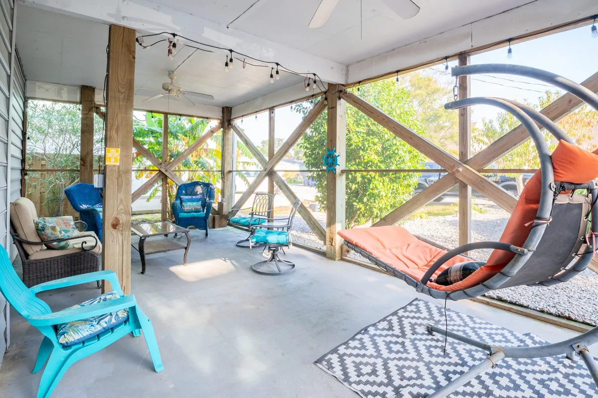 This image showcases a screened-in porch with a hanging chair, colorful chairs, a textured rug, and decorative lighting, including string lights. The arrangement creates distinct areas for both dining and casual seating, with a visible openness to the outdoors. The space is inviting and ideal for relaxation, socializing, or enjoying a coffee at sunrise.