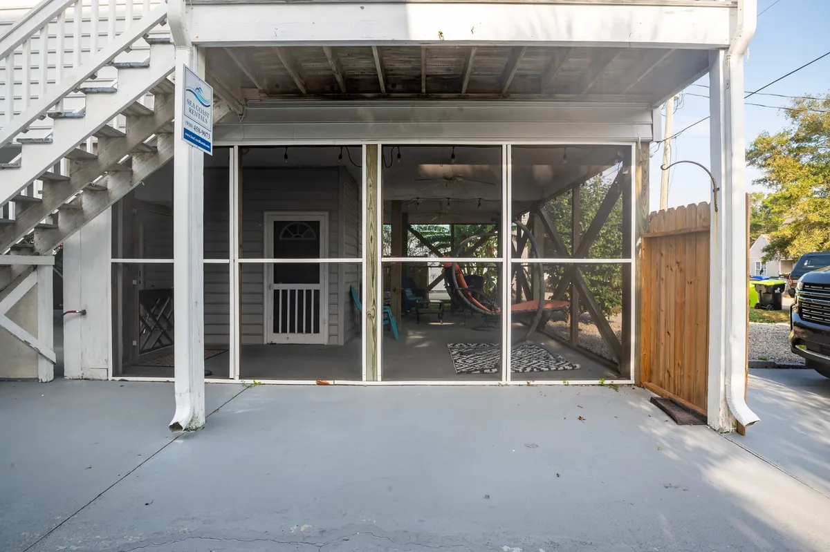 An exterior view of the screened porch area, complete with a hammock, colorful chairs, and a decorative rug. The porch is clearly sheltered yet open to the surroundings, with natural light illuminating the space and an inviting atmosphere for socializing or unwinding while enjoying the sea breeze.