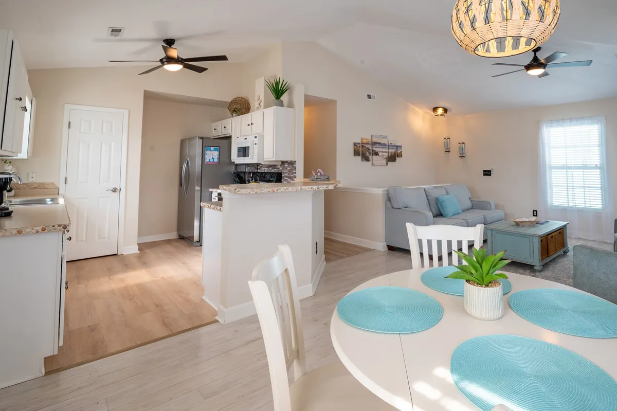 An open, sunlit interior combining a comfortable couch, a dining table adorned with blue placemats, and a visible kitchen area with modern appliances. The layout is seamless, with a light fixture above the table and a cheerful, airy atmosphere throughout.