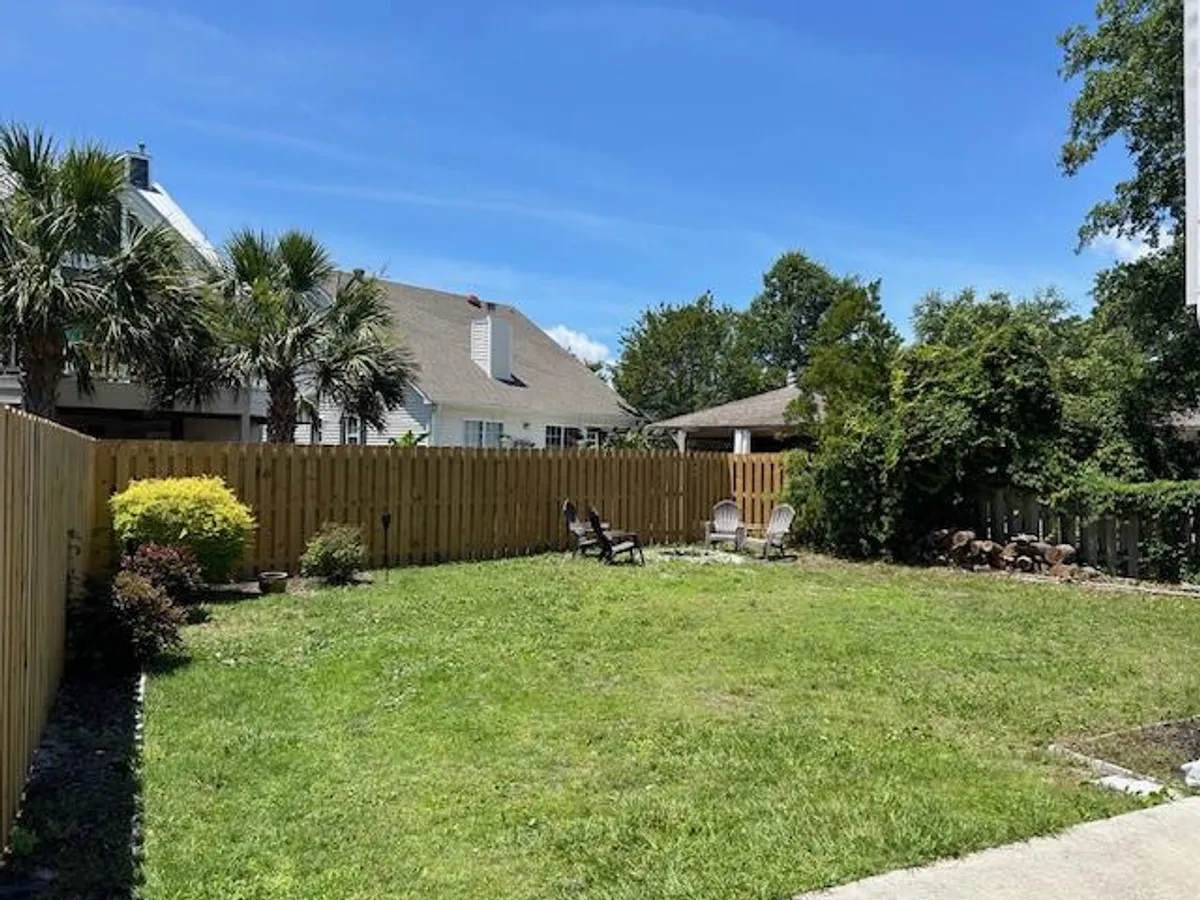 A peaceful, fenced backyard space with a lush green lawn, palm trees, and a cozy seating area with chairs. The setting invites outdoor relaxation, group gatherings, and is surrounded by a sense of privacy and tranquility, ideal for family time after a day at the beach.