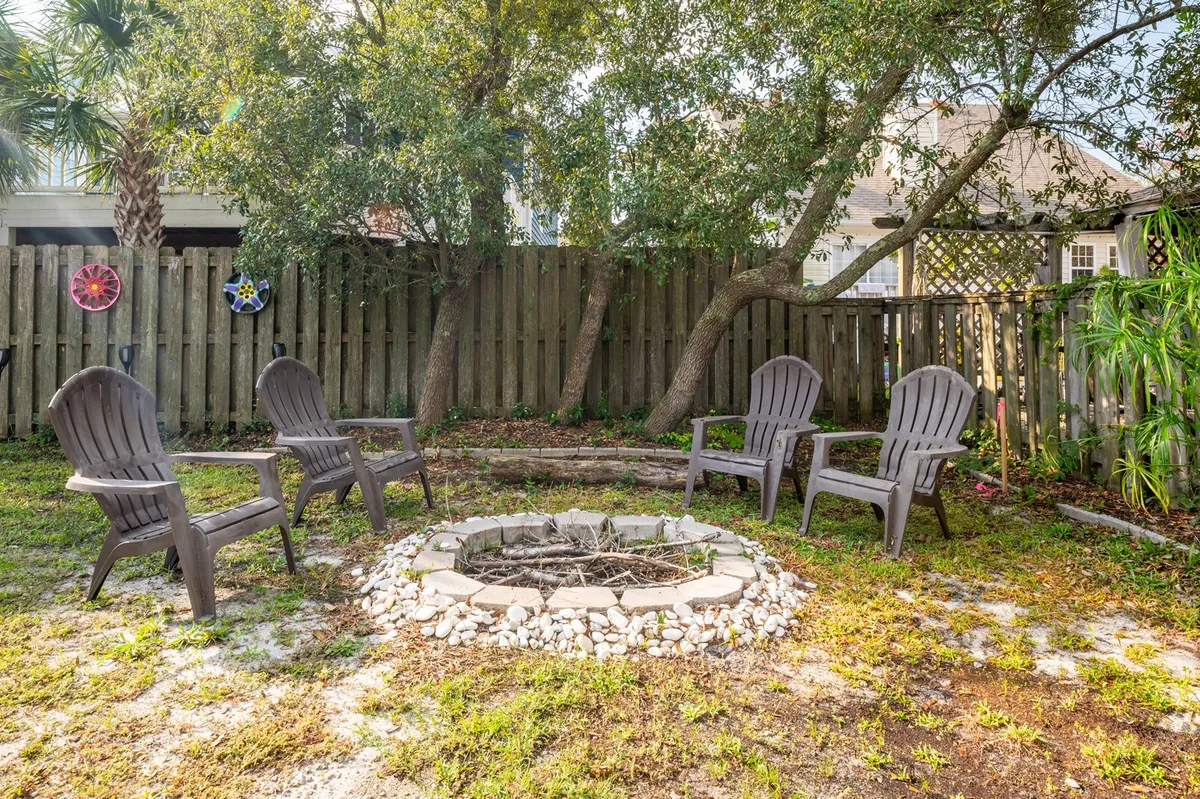 This exterior image displays a fenced backyard surrounded by trees, featuring a fire pit encircled by Adirondack chairs. The fence is clearly visible and the area exudes a cozy, communal vibe, ideal for outdoor gatherings or letting a pet explore safely under supervision.