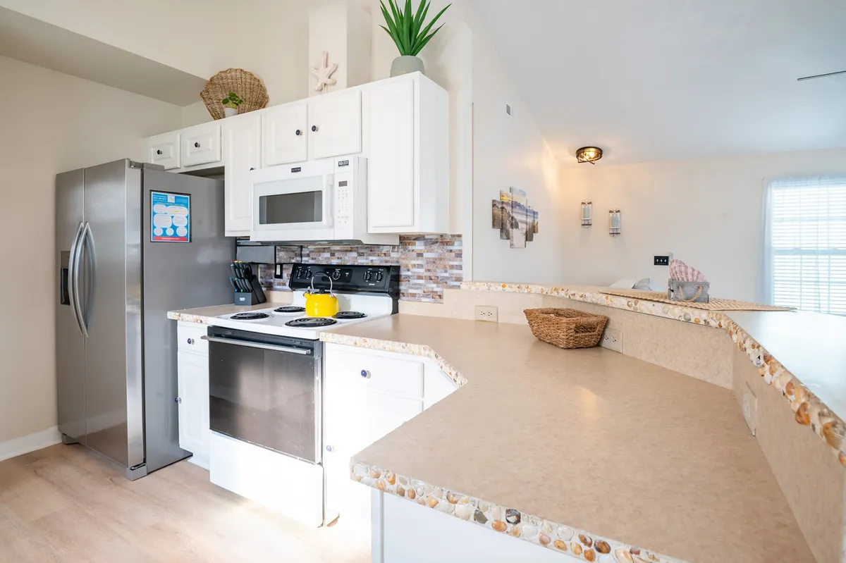 A spacious, open-concept kitchen features a stainless steel refrigerator, white cabinetry, a kitchen island with seating, and decorative plants. The area is clearly designed for group gatherings, communal dining, and entertaining, supporting both practical meals and festive occasions.