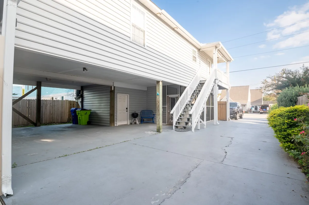 This exterior photo features the home's parking area with space for two cars, a visible staircase leading up to the main living level, and a casual seating area near a green lawn and bins. The raised structure of the home is apparent, underscoring its elevated design for coastal safety and privacy.