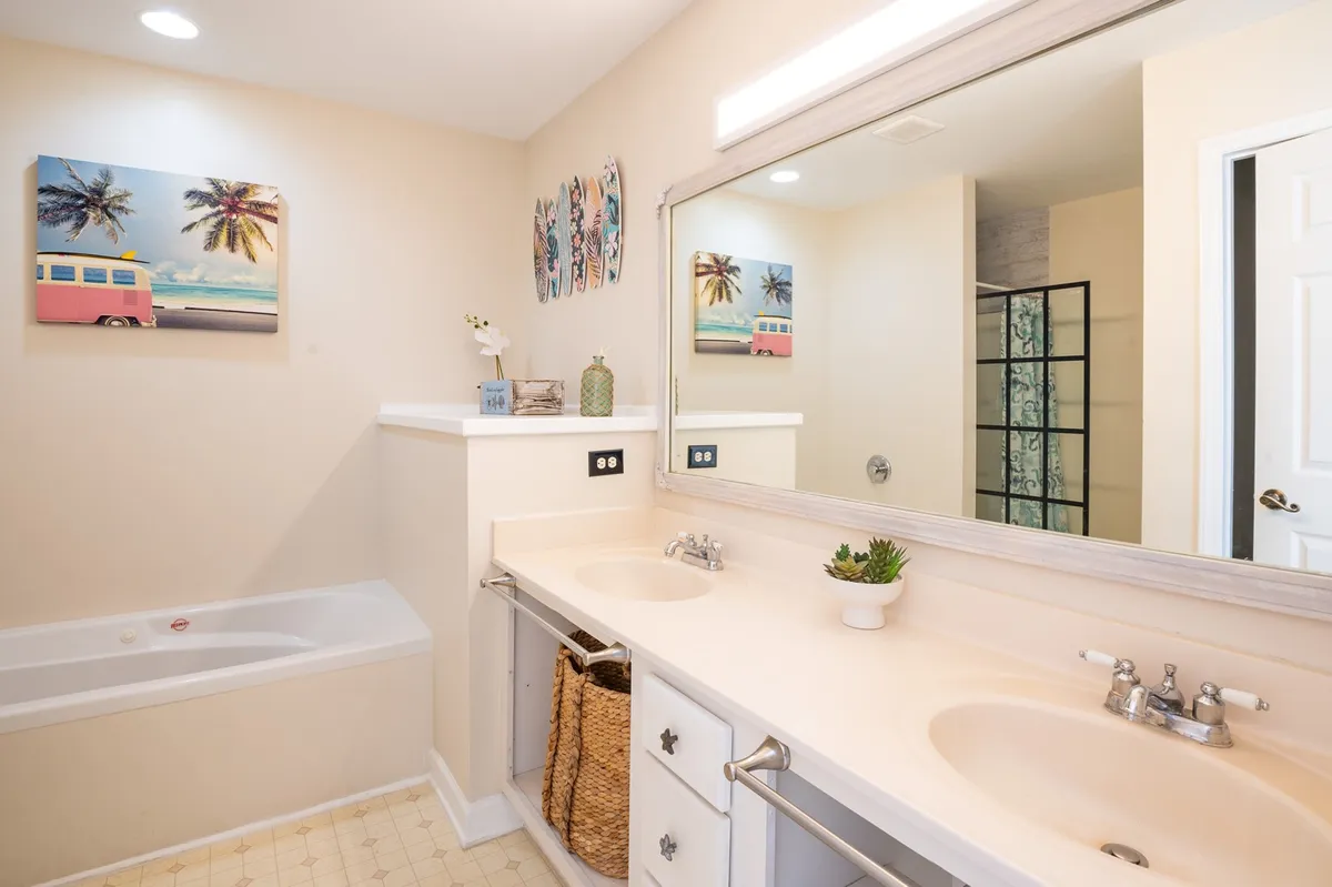 A bright and modern bathroom featuring a double vanity sink, a large framed mirror, a bathtub, and tropical-themed artwork. The space feels both refreshing and spa-inspired, ideal for unwinding.
